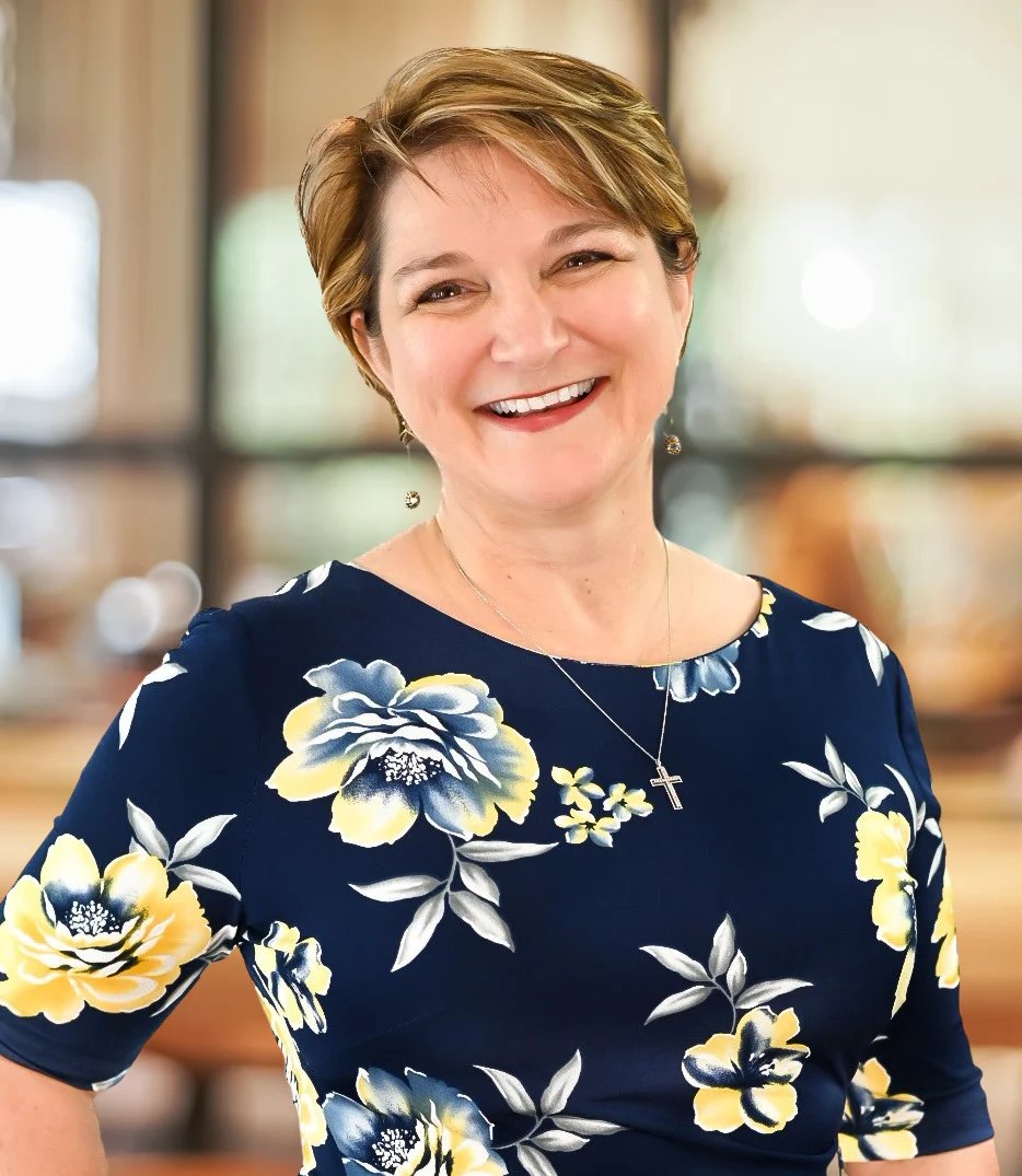 Woman with short blonde hair smiling at the camera wearing a dark navy floral dress with yellow and white flowers.